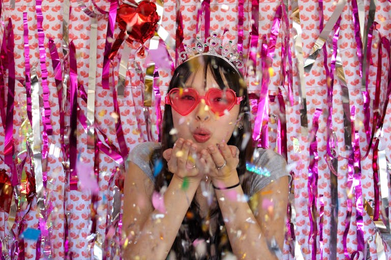Asian woman with heart-shaped glasses and tiara blowing confetti amid festive decorations.